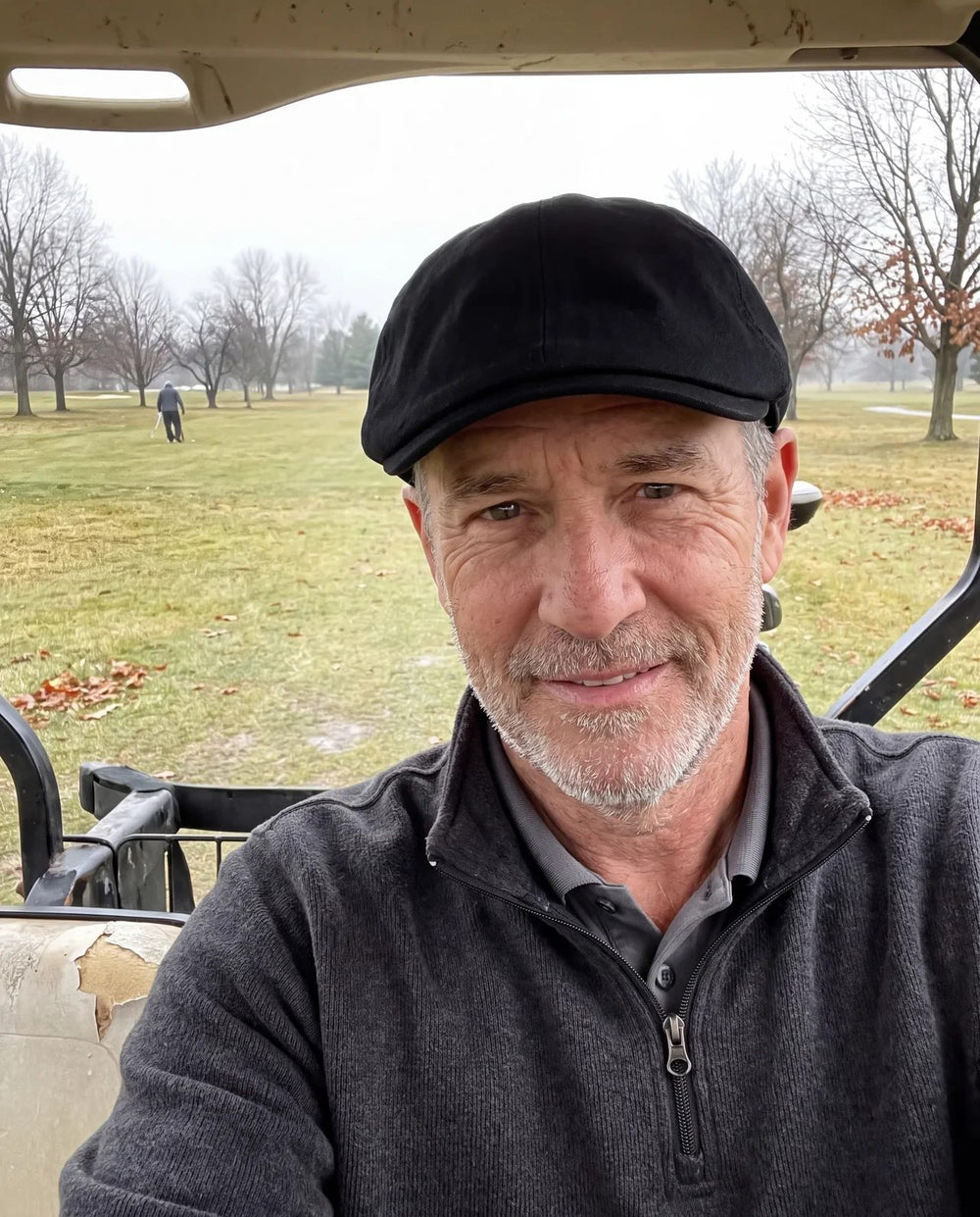 Golfer in a cart on a gray late-fall morning, black flat cap and quarter-zip